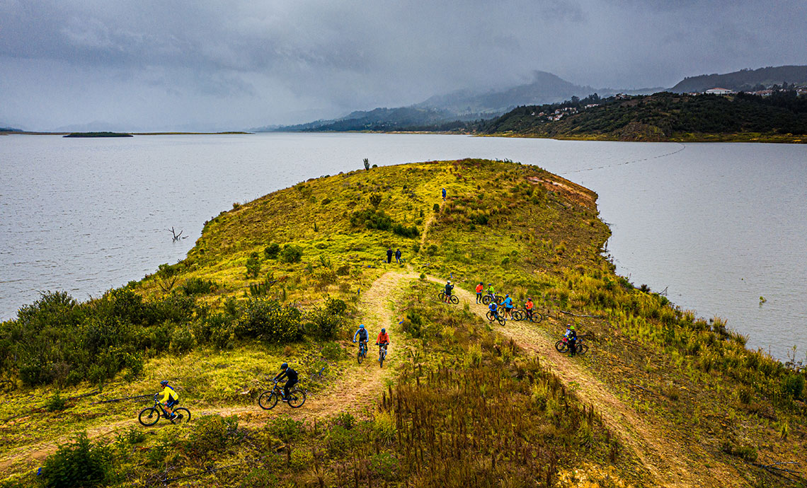 Lake surrounded by mountains; bikers ride along the edge.