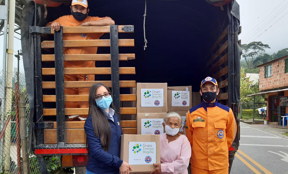 Two Civil Defense members unload humanitarian aid packages from a truck, and a GEB employee hands over a package to an older woman.