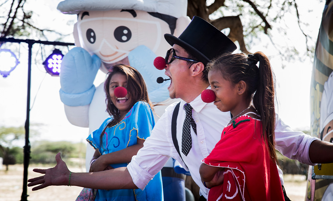 Man in white shirt, polka-dot bow tie, black hat and clown nose makes two Wayú girls, also wearing clown noses, laugh.